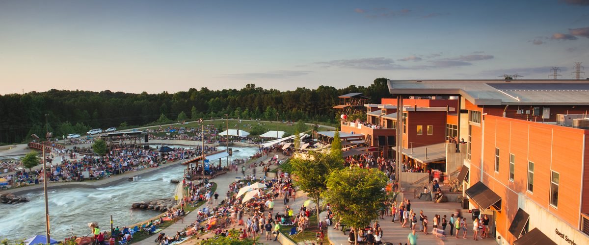 Aerial view of the U.S. National Whitewater Center at dusk, with crowds along the rapids and riverside buildings